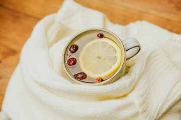 Warm cup of tea with lemon and berries, surrounded by a cozy white knitted scarf on a wooden table.