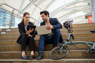 Asian business man and woman talking on stairs at train station. Using laptop and tablet, bicycle beside, urban teamwork lifestyle.