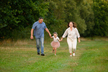 Parents with daughter are walking together in summer in meadow. Mom, dad and girl are happily booing, holding hands, laughing happily, enjoying rest together. Family, happy childhood.