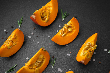 Orange pumpkin slices with seeds and rosemary on a dark surface with salt and pepper