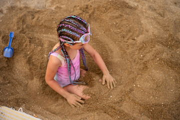 Little girl with colorful braids wearing swimming goggles and a pink swimsuit playing in the sand...