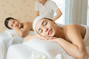Two women enjoying relaxing spa massage side by side on white beds, eyes closed and smiling peacefully, representing wellness, beauty care, and relaxation in luxurious spa setting.