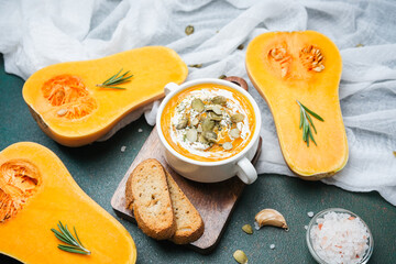 Butternut squash soup with cream and pumpkin seeds, served with bread slices and herbs on a table.