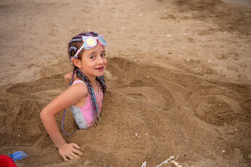 Little girl with colorful braids and swimming goggles, buried in the warm sand, smiling joyfully...