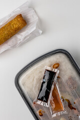 Asian takeout meal in plastic container with sauce packets and crispy egg roll on white background