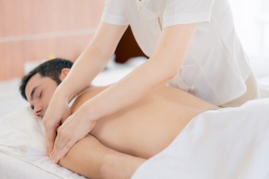 A man lying on his stomach receiving a professional shoulder massage at a spa. The therapist uses a gentle palm technique to apply pressure, muscle relaxation and stress relief in a environment