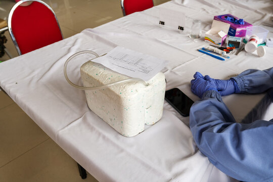 Medical staff with gloves sitting beside styrofoam cooler box, healthcare tools, and documents on table, showing preparation for blood donation, vaccine storage, or clinical service.