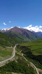 A winding road snakes through a lush valley nestled amongst towering snow-capped mountains under a vibrant blue sky.