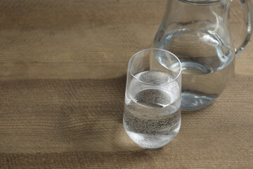 Glass of water with jug at wooden table, closeup. Drinking clear water in the morning for health. Space for text 
