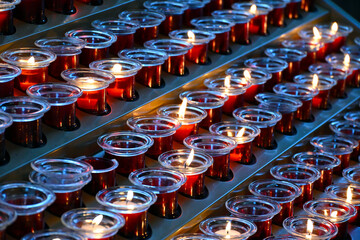 Fototapeta premium Small votive candles, lighted and placed in red glass candlesticks among empty ones, interior details of a catholic church