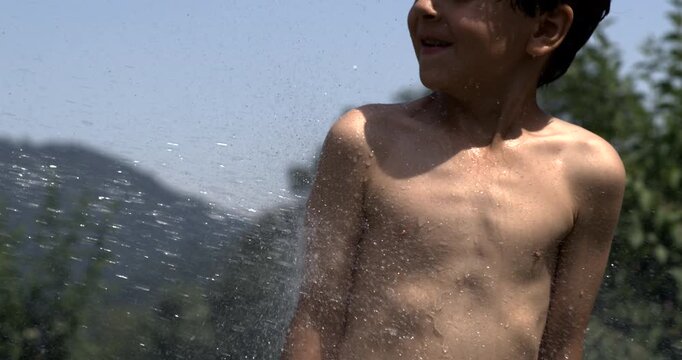 Wet torso of young boy with water spraying across chest in sunlight, wearing swim shorts, captured in super slow motion outdoors