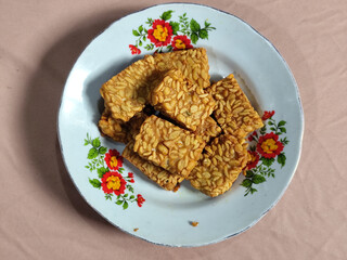 A pile of crispy fried tempeh that looks delicious and tasty, in a glass plate with a floral motif.