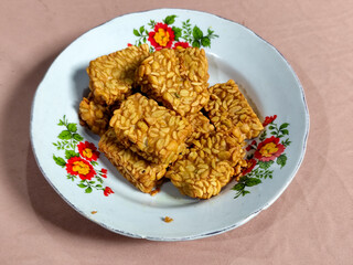 A pile of crispy fried tempeh that looks delicious and tasty, in a glass plate with a floral motif.