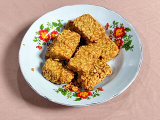 A pile of crispy fried tempeh that looks delicious and tasty, in a glass plate with a floral motif.