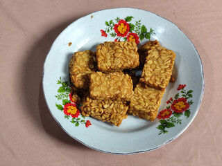 A pile of crispy fried tempeh that looks delicious and tasty, in a glass plate with a floral motif.