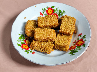 A pile of crispy fried tempeh that looks delicious and tasty, in a glass plate with a floral motif.