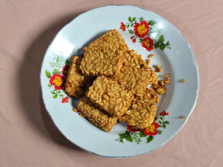 A pile of crispy fried tempeh that looks delicious and tasty, in a glass plate with a floral motif.