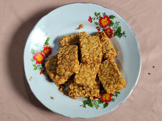 A pile of crispy fried tempeh that looks delicious and tasty, in a glass plate with a floral motif.