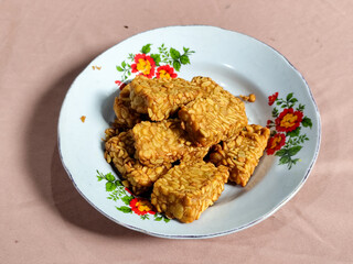 A pile of crispy fried tempeh that looks delicious and tasty, in a glass plate with a floral motif.