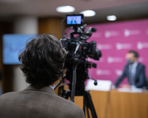 Person with curly hair is seen from behind, operating video camera during press conference. setting features professional backdrop with signage, indicating formal event