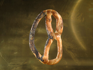 A single pretzel, coated with poppy seeds, hovers against a blurred gold background. The pastry is...