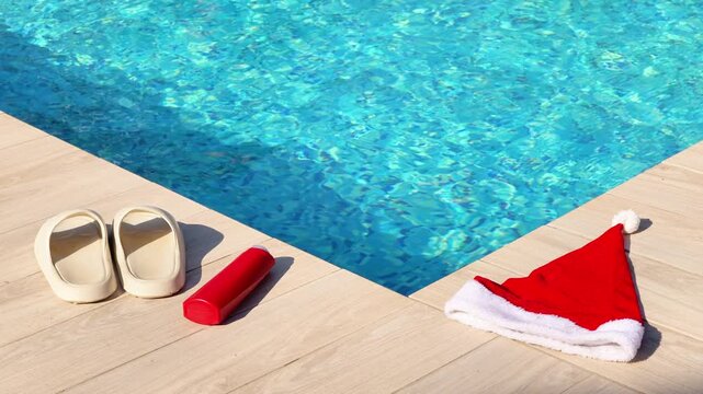 A Santa hat and poolside essentials like sandals and a water bottle sit on a wooden deck next to a vibrant blue swimming pool.