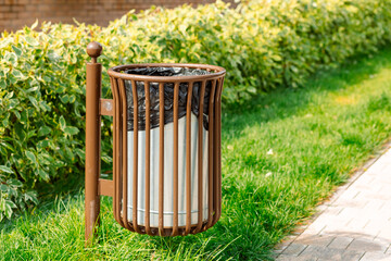 A street trash can made of metal with vertical bars. It is installed on green grass next to a path. In the background, bushes with yellow flowers and a brick wall are visible.