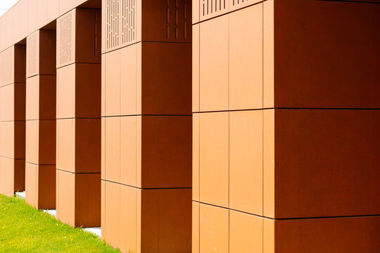 A modern facade made of terracotta panels with a geometric pattern. Bright sunlight highlights the texture and color of the material. At the base is lush green grass.