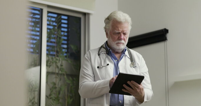 Elderly Doctor Standing Alone While Reviewing Patient Information on Tablet in Modern Clinical Office Setting