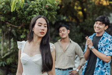 A reserved young Asian woman looks downward in quiet discomfort while two men behind her leer and catcall with smug expressions in a garden setting.