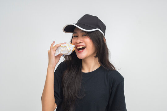 A playful young Asian woman in a half body shot smiling with her mouth open, about to take a bite of a burger, teasingly inviting the camera. Isolated on a white background.