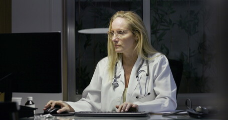 Female doctor taking notes while working late at desk, glancing away from computer monitor with...