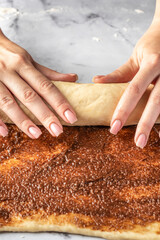 Women's hands roll the dough into a roll on white marble background. Making cinnamon rolls process. Cinnabon preparation