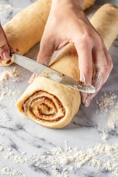 Women's hands cutting a roll of dough on white marble background. Making cinnamon rolls process. Cinnabon preparation
