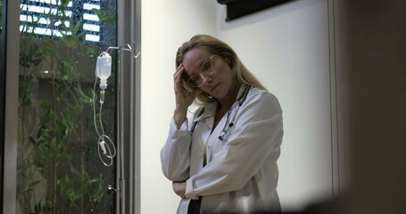 Female doctor looking down with tired expression, resting head in hand while standing beside IV...
