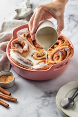 Female hands pouring glaze over freshly baked cinnabon buns in a ceramic baking dish, white marble background. Making cinnamon rolls process