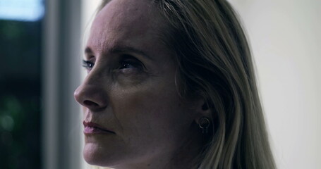 Middle aged woman gazing into distance with thoughtful expression in softly lit indoor setting,...
