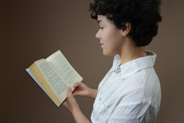Young woman with short curly hair, reading a book. Horizontal studio shot with copy space on brown background. Ideal for education, study, literature and library.