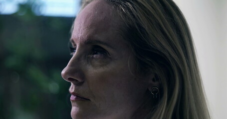 Middle aged woman gazing into distance with thoughtful expression in softly lit indoor setting,...
