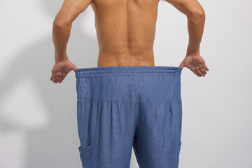 
A fit man with a toned physique shows off his significant weight loss by wearing his old, oversized, and loose denim trousers.