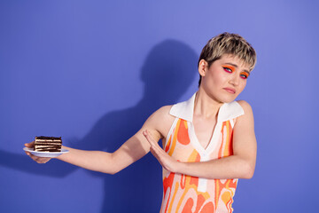 Young woman with vibrant makeup refusing a slice of chocolate cake in front of a vivid purple...