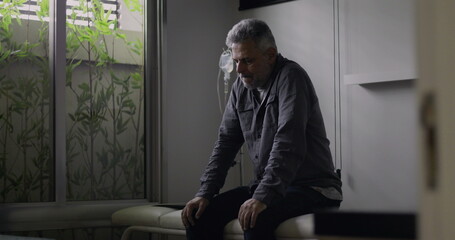 Man sitting alone in clinical room with IV drip behind, looking down in silence, experiencing...