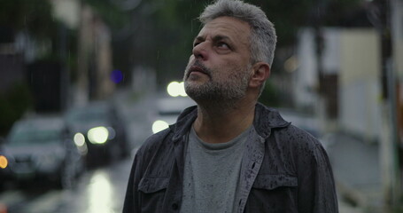 Middle aged man standing in rain with distant look, wet clothes and somber expression, surrounded...