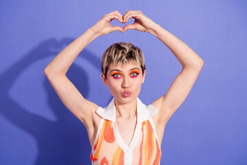 Joyful young woman forming a heart shape with hands, embracing vibrant makeup and a bold retro-style outfit against a purple background