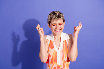 Excited woman with short hair crossing fingers and smiling joyfully against a vibrant purple...