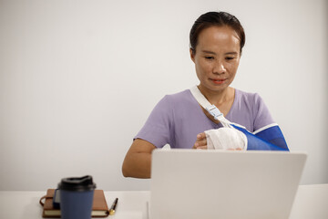 A person in a purple shirt sits at a desk with their right arm in a blue mesh sling, typing on a laptop with their left hand, showing resilience, adaptability, and determination.