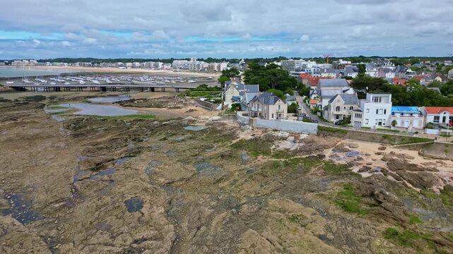 Reverse aerial drone shot over coastal rocks near Pornichet, pulling back to reveal the marina, Libraires Beach, and the cityscape under a partly cloudy sky - Brittany France