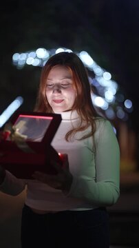 Woman opening glowing Christmas gift box at night with festive lights background