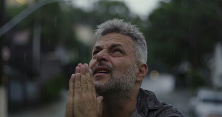 Middle aged man looking upward with raised hands in rain, emotional expression of questioning or...