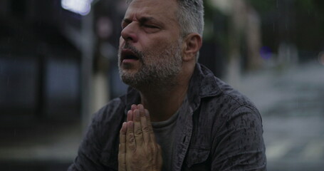 Middle aged man with eyes shut and hands clasped in desperate prayer or emotional plea, standing in...